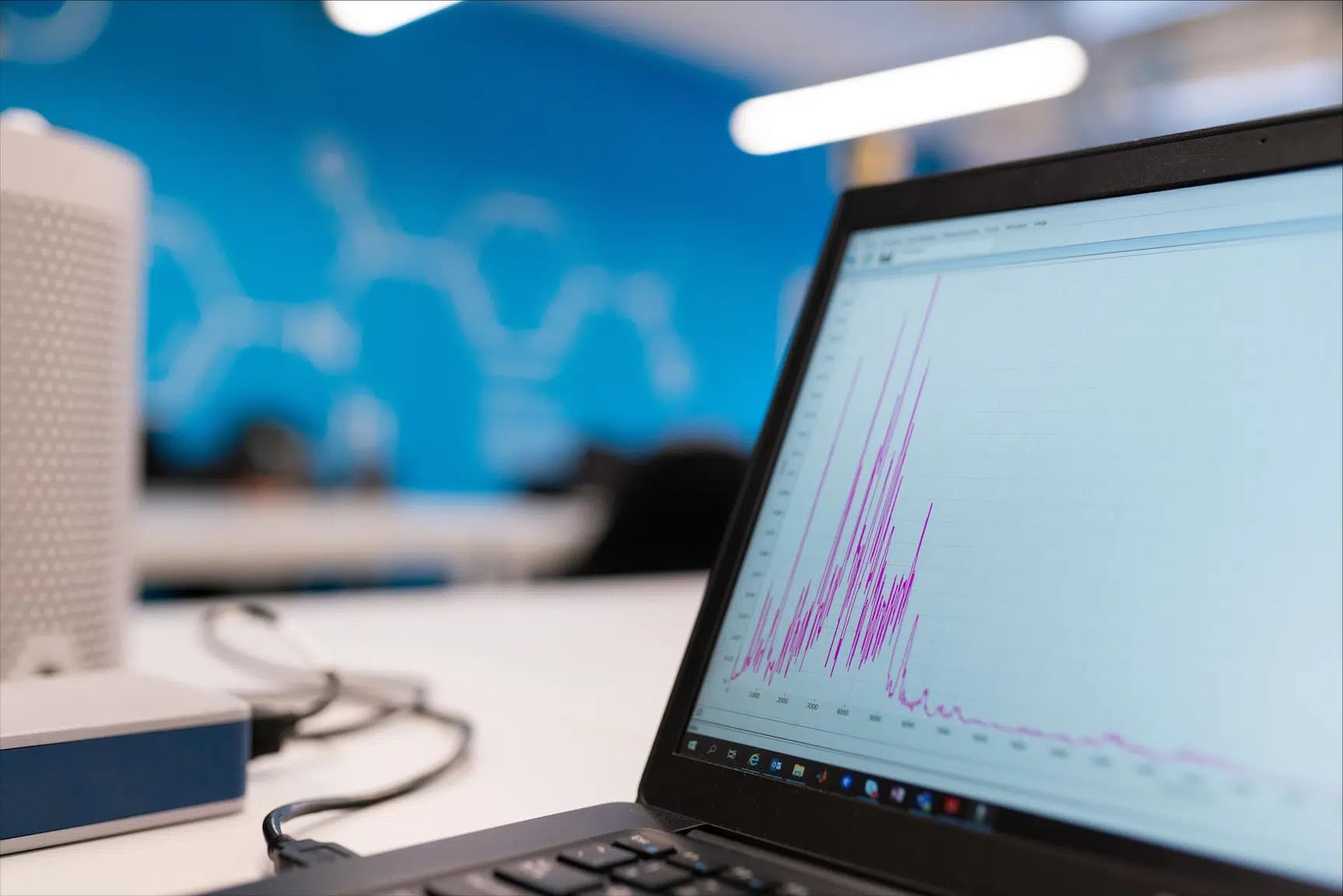 A laptop on a desk shows a chart with sharp peaks; behind it, a blue wall with a data visualisation.