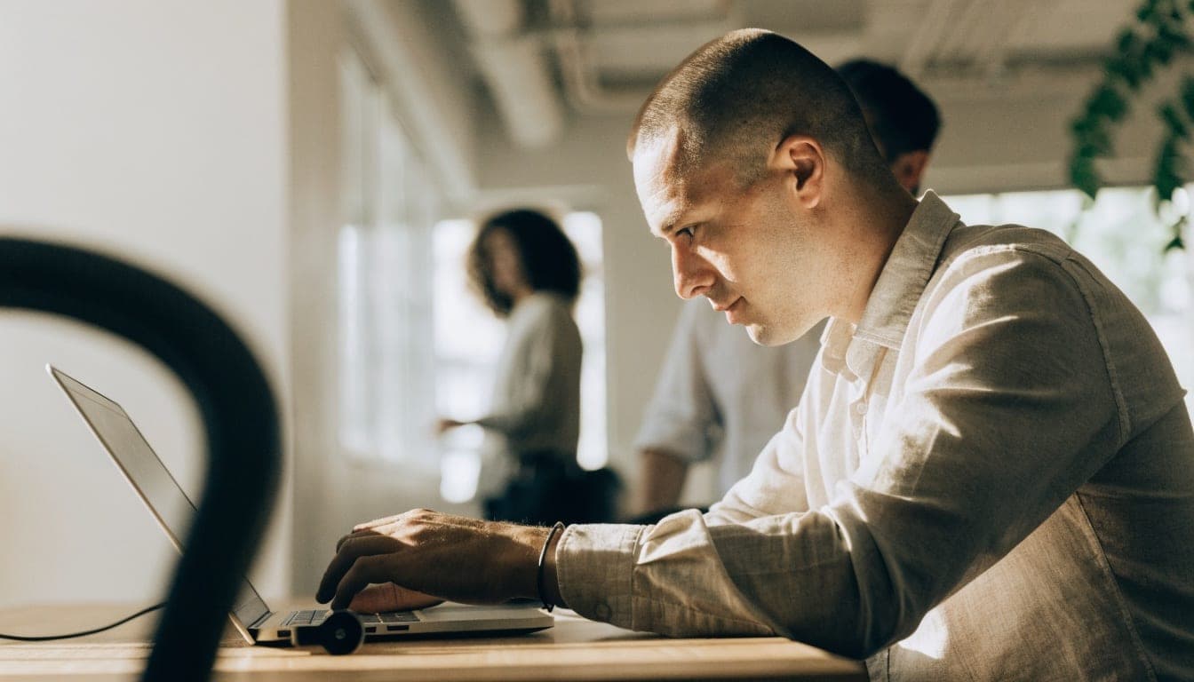 Man working with focus on a laptop in an office with colleagues in the background