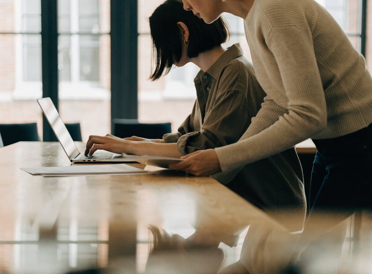 A content marketer typing a request to an AI assistant on her laptop while a colleague looks on