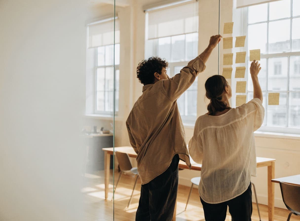 Two people arranging sticky notes on a glass wall to prioritise the features of a first product version