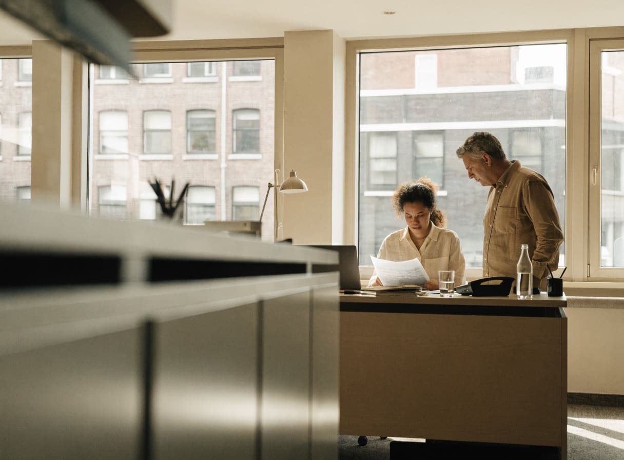 An employee reading a document at her desk next to a laptop while a colleague looks on