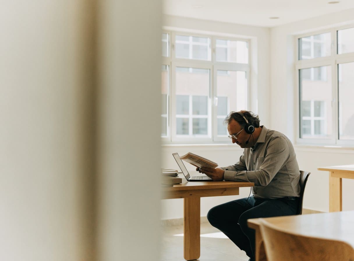 An employee wearing a headset reads a document at a desk with a laptop