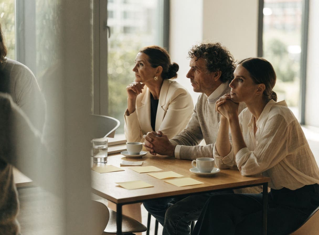 Three colleagues at a table with coffee and yellow sticky notes, listening to an explanation